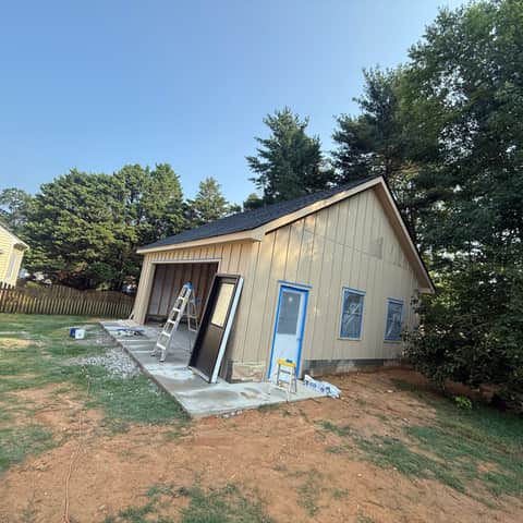 The primed board-and-batten garage with its windows masked in blue tape ready for paint, and an entry door leaning against the wall