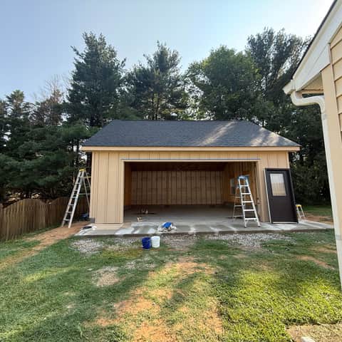 The detached garage in primer with its bay open, showing the interior framing and sheathing, with stepladders and paint buckets on the slab