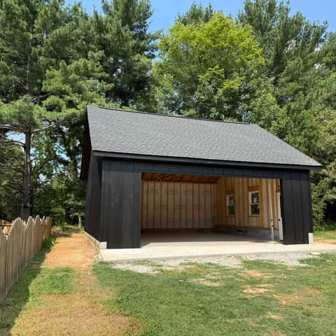 A finished detached garage with black board-and-batten siding and a new shingle roof, its bay open to show the framing and sheathing inside